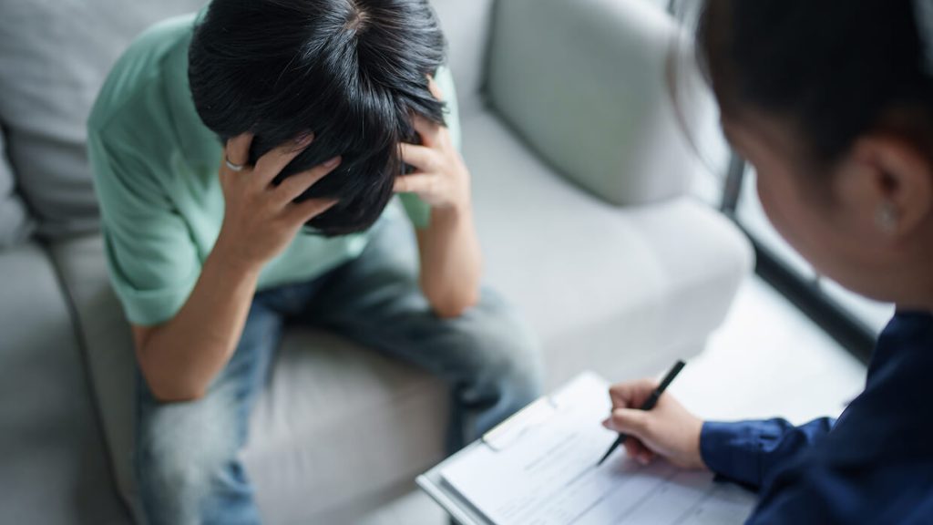 A child sitting on a couch with their hands on their head, appearing distressed, while an adult writes notes on a clipboard nearby.
